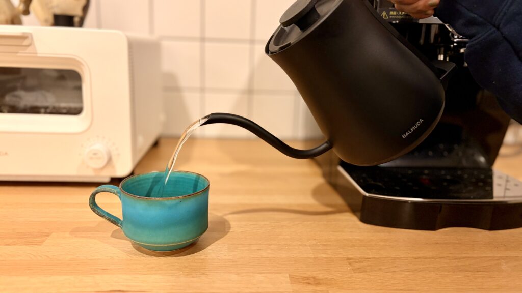 BALMUDA The Potの黒い電気ケトルからお湯が緑のマグカップに注がれているシーン。背景には白いトースターとカウンターが見える。